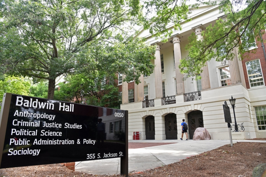 Exterior view of Baldwin Hall at the University of Georgia, with large white columns and arched doorways. A black sign in the foreground lists departments housed in the building, including Anthropology, Criminal Justice Studies, Political Science, Public Administration & Policy, and Sociology. A student walks toward the entrance.