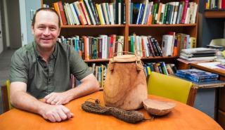 Bram Tucker sits in his office smiling at the camera. There is a full bookshelf behind him, and artifacts on the table in front of him.