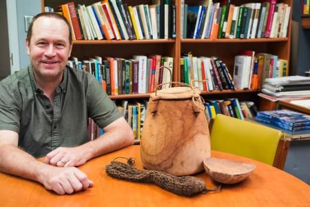 Bram Tucker sits in his office smiling at the camera. There is a full bookshelf behind him, and artifacts on the table in front of him.