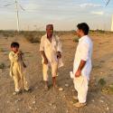 Three people—an elderly man, a young boy, and another adult man—stand in conversation on a dry, open landscape scattered with low shrubs. They wear traditional light-colored clothing and hold wooden walking sticks. In the background, multiple large wind turbines rise across the horizon under a partly cloudy sky, suggesting a juxtaposition of local community life and renewable energy development. A few goats can be seen grazing in the distance.