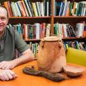 Bram Tucker sits in his office smiling at the camera. There is a full bookshelf behind him, and artifacts on the table in front of him.